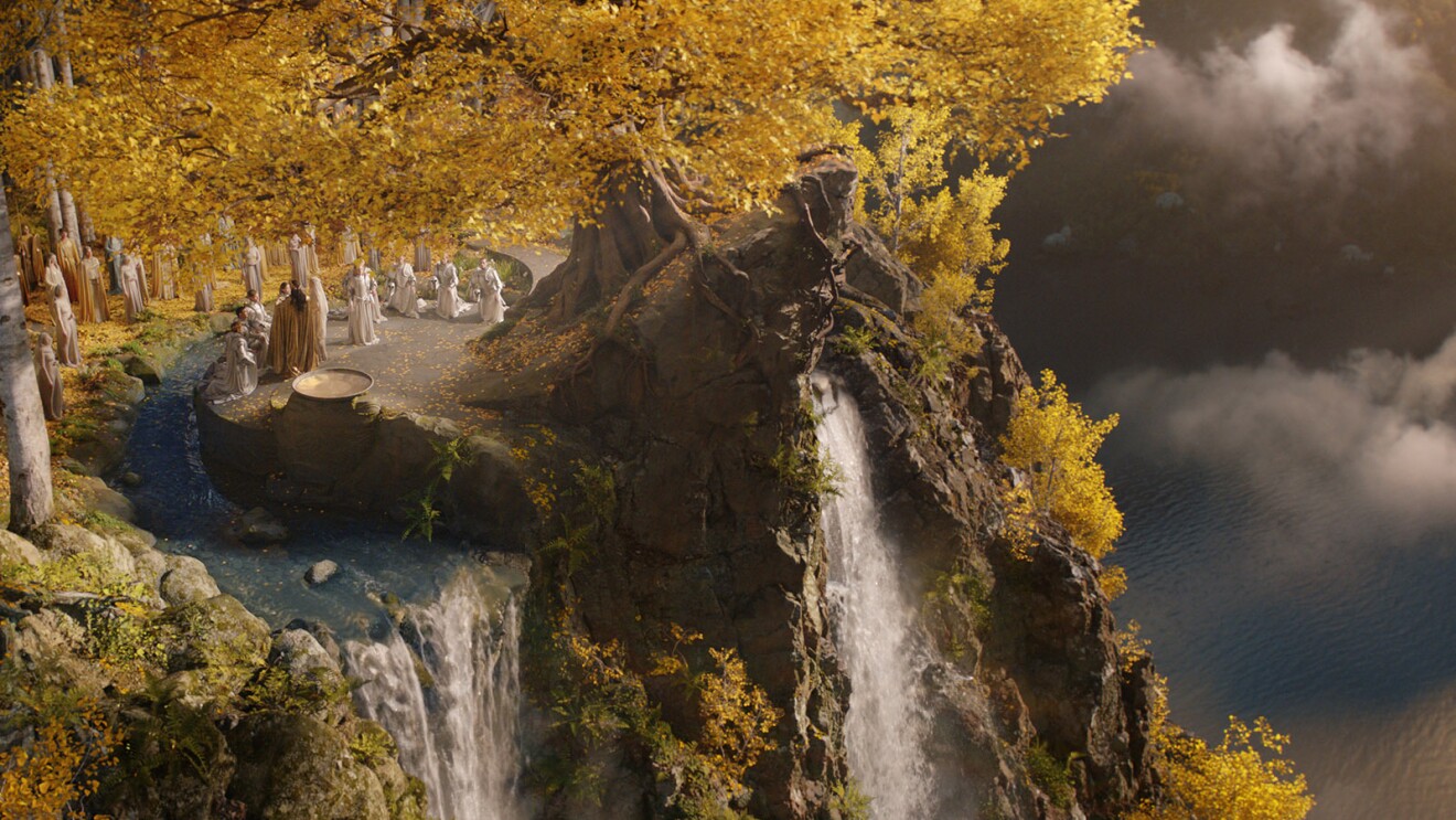 An image of a cliff with a waterfall on the ledge and a large body of water below. There are treed in the background and people gathered around the edge. It is a scene from Amazon's The Lord of the Rings: The Rings of Power series.