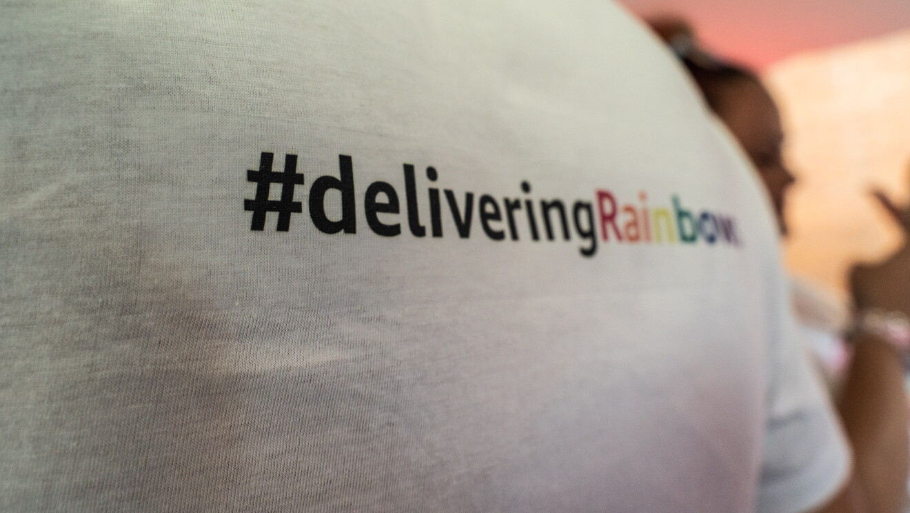 Camiseta de glamazon para Día internacional del orgullo LGTBI en Madrid y Barcelona. La camiseta tiene el mensaje delivering rainbows inspirada en el lema de Amazon delivering smiles. La camiseta es de color blanco y las letras están en negro menos rainbows que cada letra es de un color diferente imitando el arcoiris.