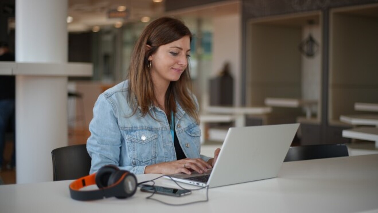 A young woman is working at her laptop. She has a blue shirt and a pair of orange headphones are lying on a table.