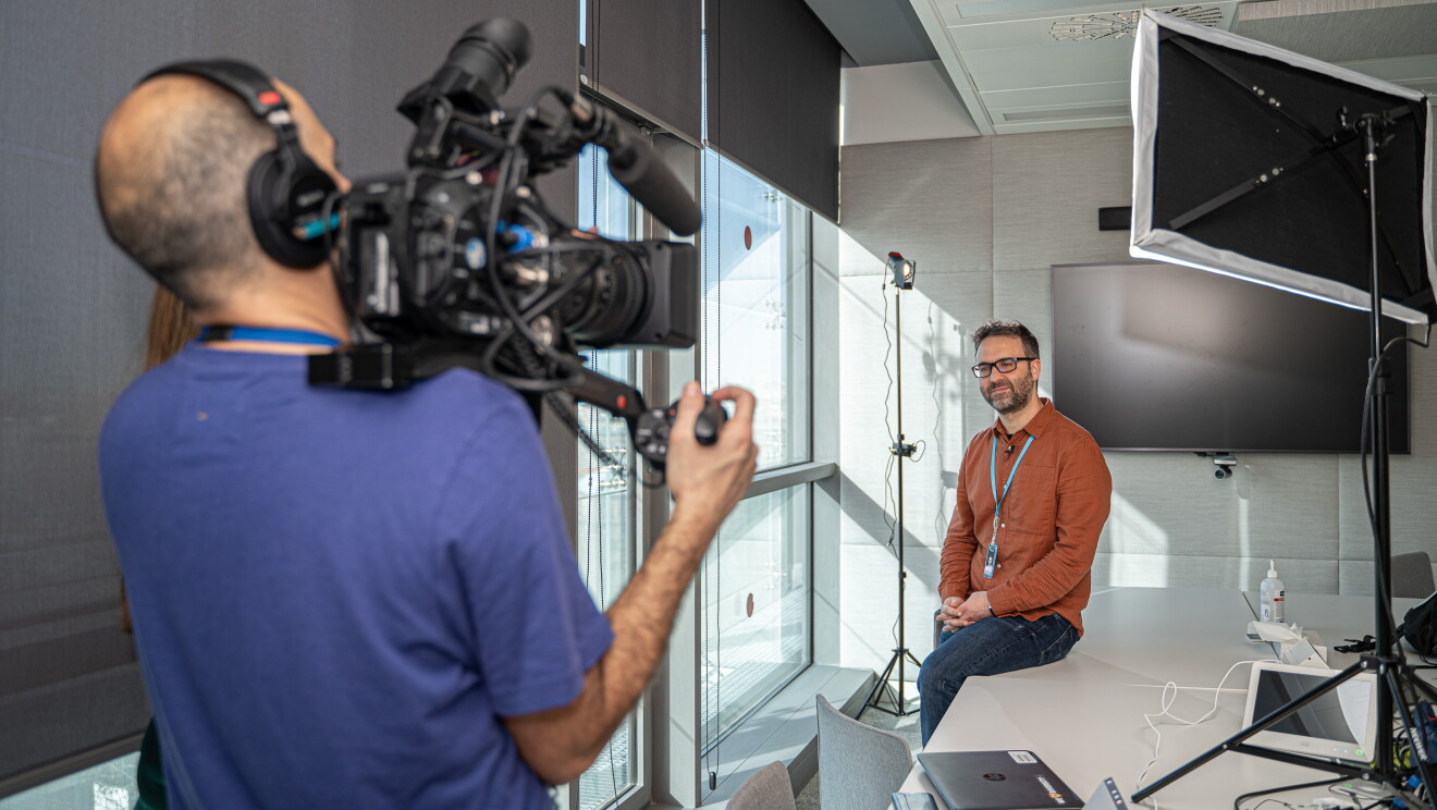 Daniel Méndez, Content Program Manager de Alexa, en una sala de trabajo de las oficinas de Amazon en un momento de la grabación. Daniel aparece en el fondo con una camisa de color marrón. Las ventanas están en el lado izquierdo. En primer plano y desenfocado aparece el cámara con una camiseta azul de manga corta.