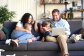 A family of four smiles and sits on a couch looking at a laptop.