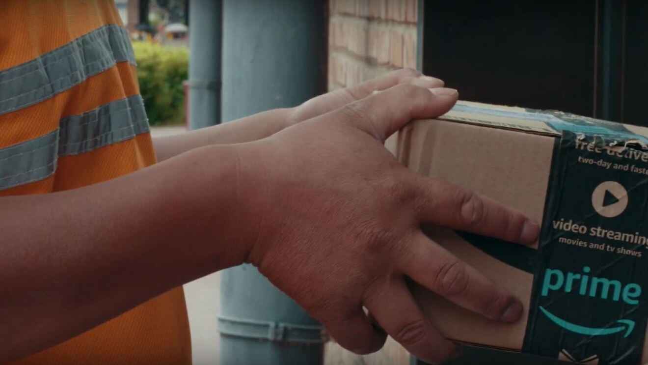 An Amazon delivery driver hands over a package to a customer. The box tape features the smiling Prime logo and video streaming icon.