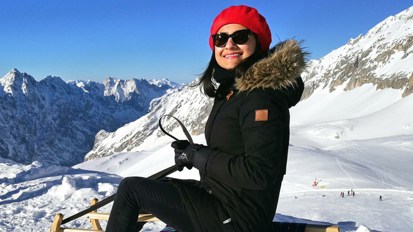 Mehak in den Alpen beim Schlitten fahren. Sie trägt eine rote Pudelmütze und eine Sonnenbrille. Im Hintergrund sieht man die schneebedeckten Alpen.