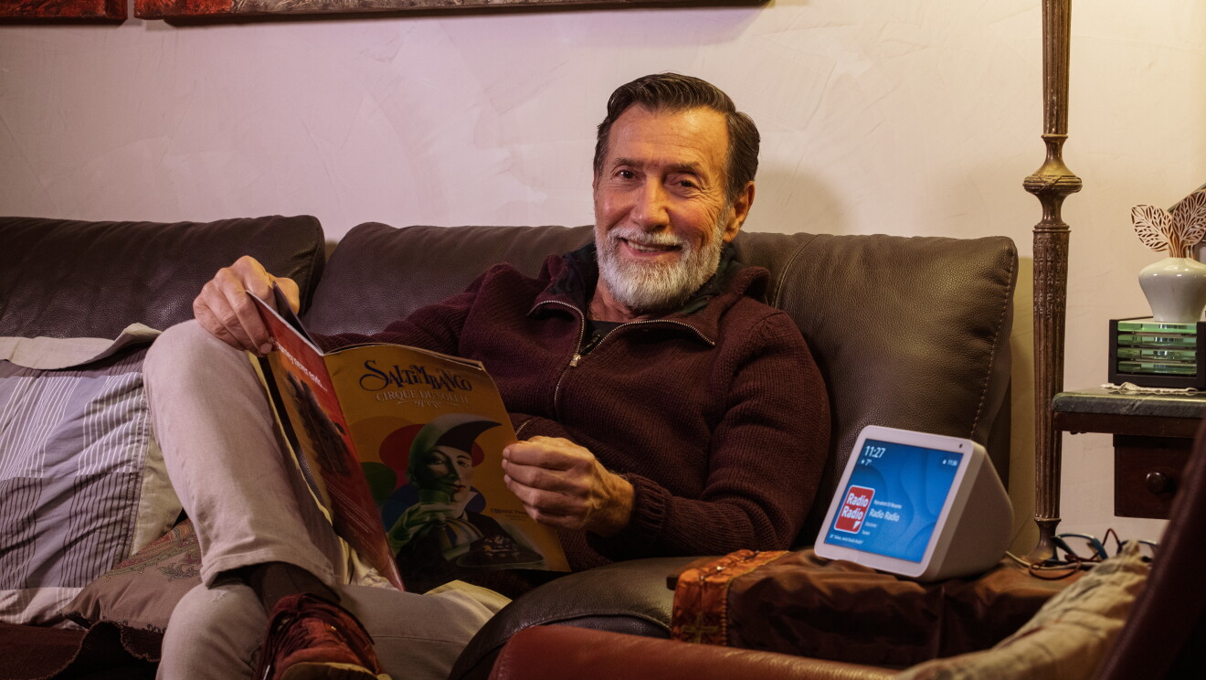 Un uomo di mezza età è rivolto verso un device Echo Show, che ha lo schermo acceso. L'uomo è al centro dell'immagine, seduto su un divano in pelle marrone. Il device Echo, a destra, è poggiato su un tavolino. 