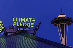 An image of the top of Climate Pledge Arena in Seattle at night. There is the illuminated neon sign that says "Climate Pledge Arena" on top of the stadium and the Space Needle in the background.