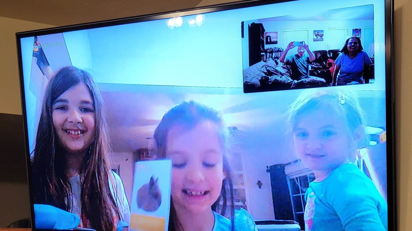 An image of a television screen with three children smiling on a video call with their family.