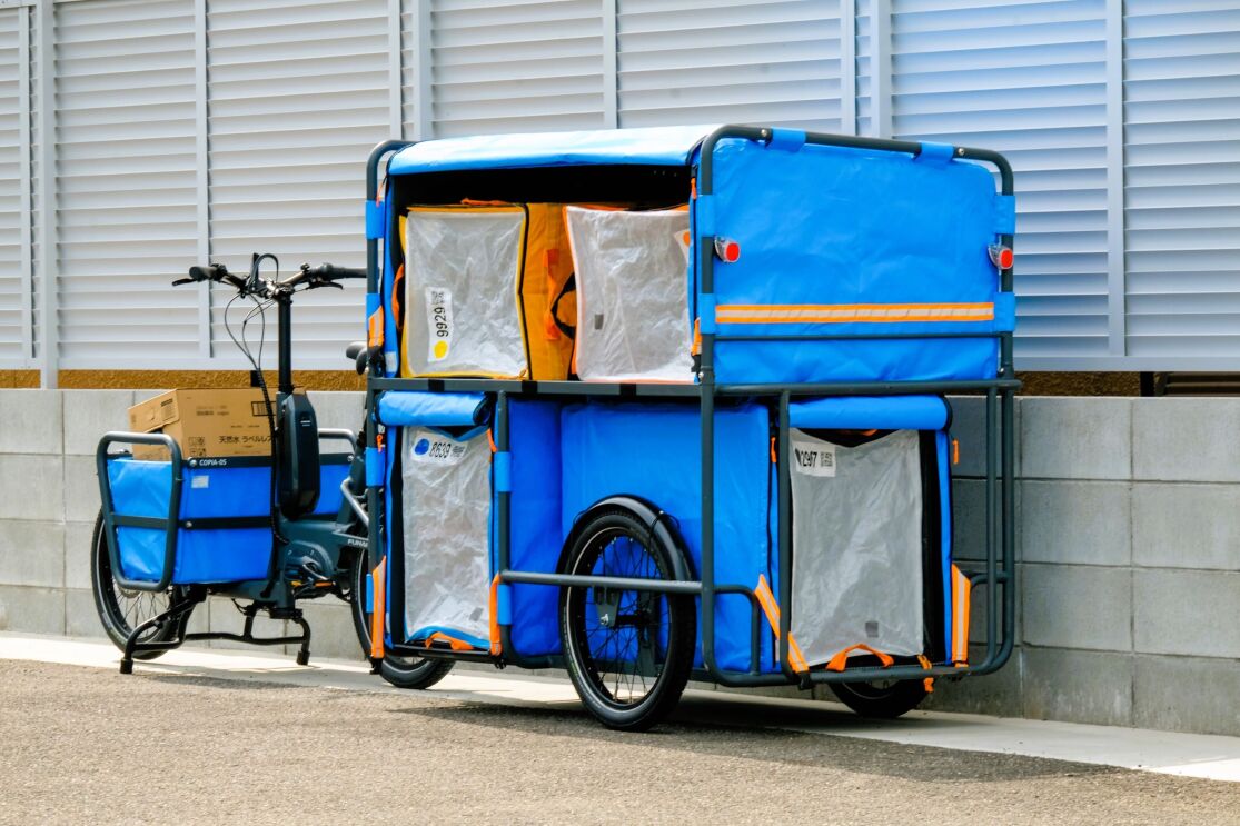 屋外の塀の前にリヤカーと前輪部分にカゴがとり付けられた電動アシスト自転車が置かれている。リヤカーには大きな青いカバーが付けられた箱が積まれている