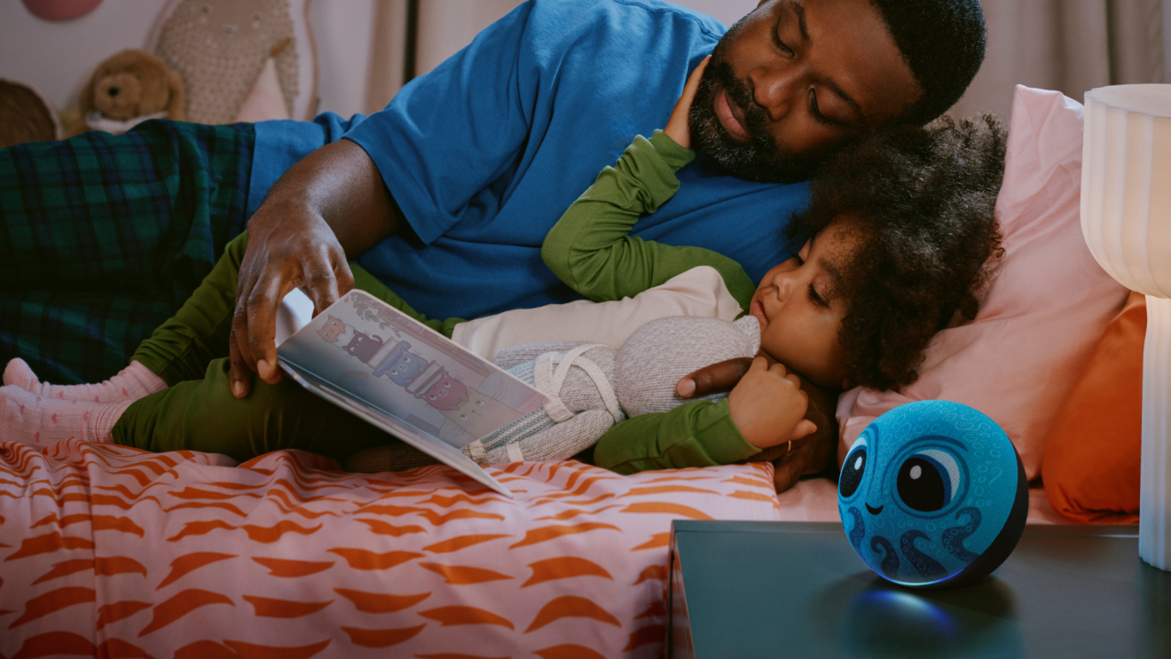 A parent and child laying on a bed, looking at a book. Beside them is an Amazon Kids Echo Dot placed on a nightstand.