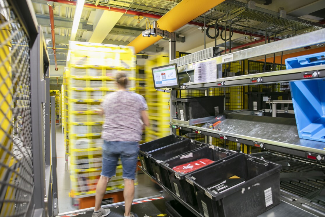 eine Mitarbeiterin befüllt ein gelbes Regal mit Artikel, Unter dem Regal sieht man den Roboter.