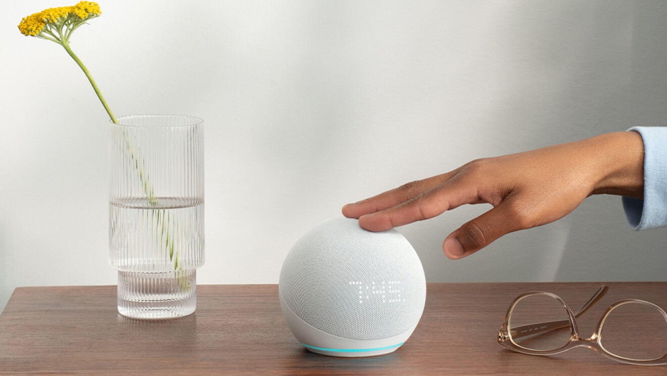 Sobre uma mesa, um Echo Dot branco sendo tocado por uma mão. Há também um óculos e um copo com uma flor amarela