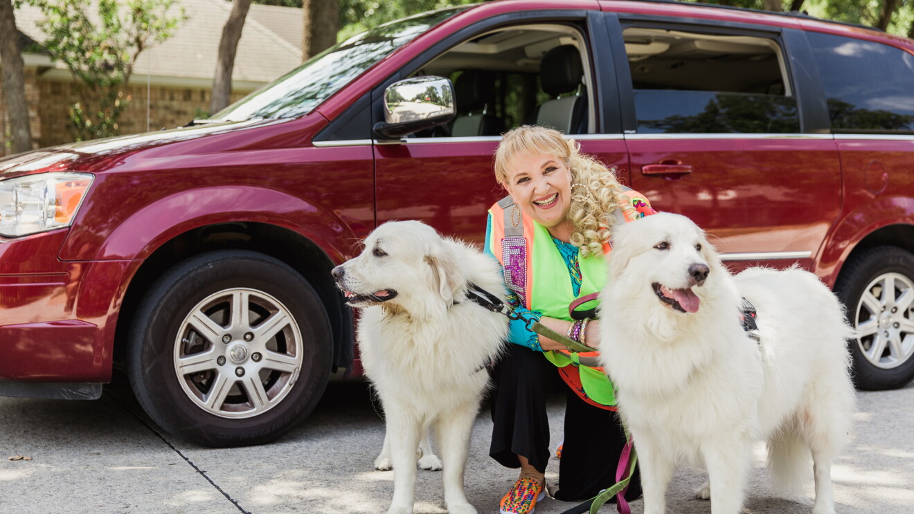 A woman in a bedazzled Amazon Flex vest stands in front of her maroon mini-van. She is smiling at the camera and standing with her two white dogs, who are wearing service animal vests. 