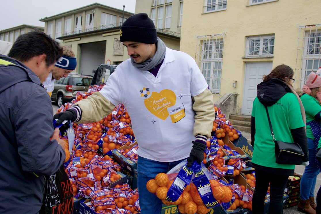 Ein Volunteer von Amazon, gekleidet im weißen Shirt mit Aufdruck "Amazon Volunteers", gibt Orangen an Bedürftige aus. 