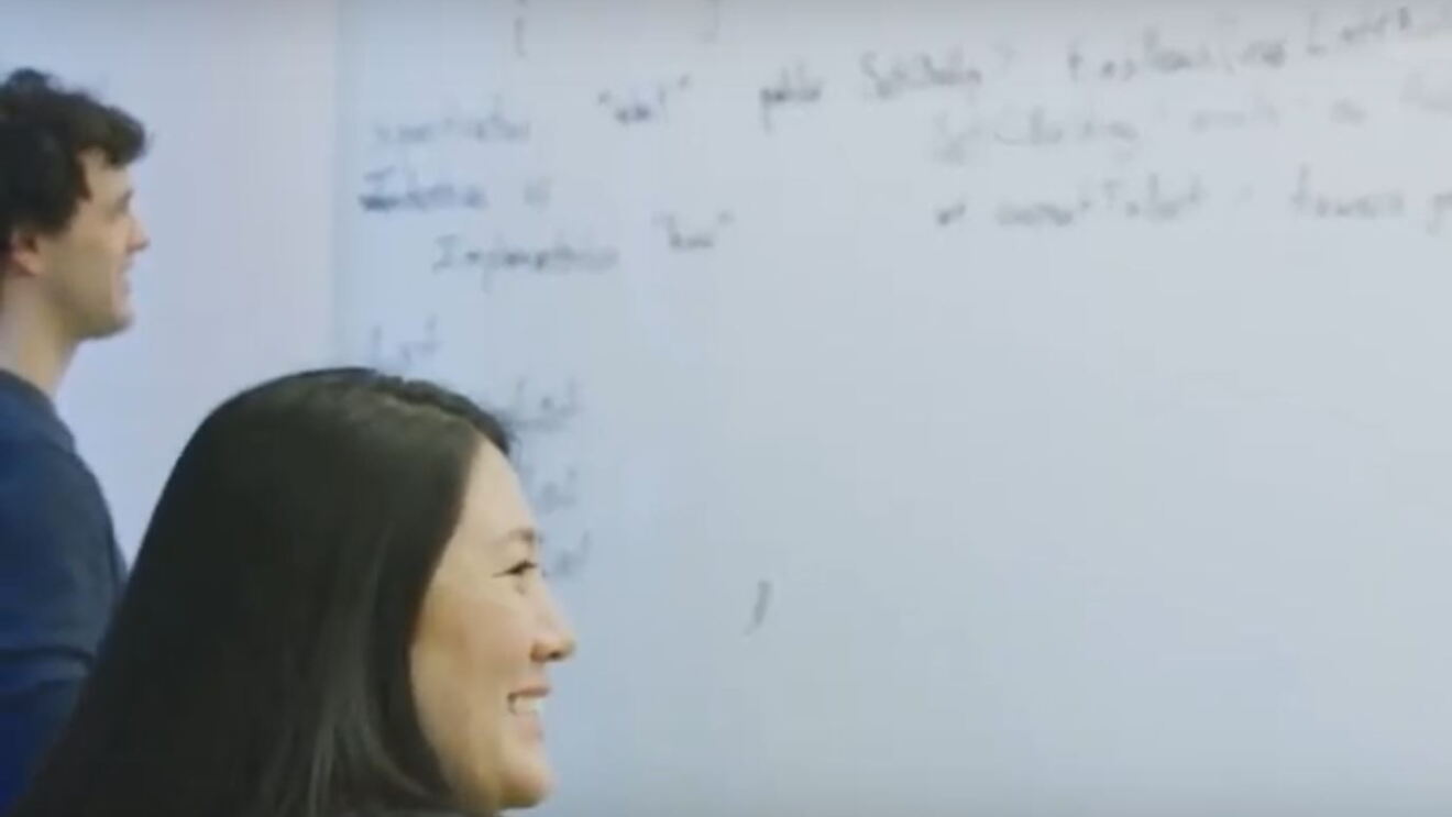 A woman in a classroom setting smiles as she looks to her right. Behind her, a man writes on a white board.
