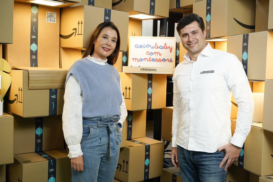 En un escenario formado por cajas de Amazon de cartón. Marga López y Lucas Díez son los fundadores de AEIOU Baby, una pyme de Talavera de la Reina (Toledo) que, desde que nació en 2016, vende productos exclusivamente en Amazon. Marga va con unos jeans azules de tiro alto con cinturón de tela, atado en un lazo en la cintura. Debajo una camisa y un jersey sin mangas azul. Lucas lleva una camisa blanca con el nombre de la empresa AEIOU Baby. Lleva el pelo corto, jeans i las mano en apoyados en los bolsillos. Entre ellos y apoyados en una caja de Amazon, la pizarra de madera con letras azules y rojas y el texto aeioubaby.com Montessori. 