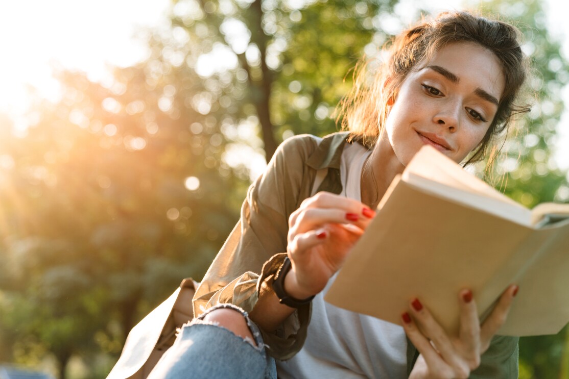 Image of young woman smiling and reading book in green park_1