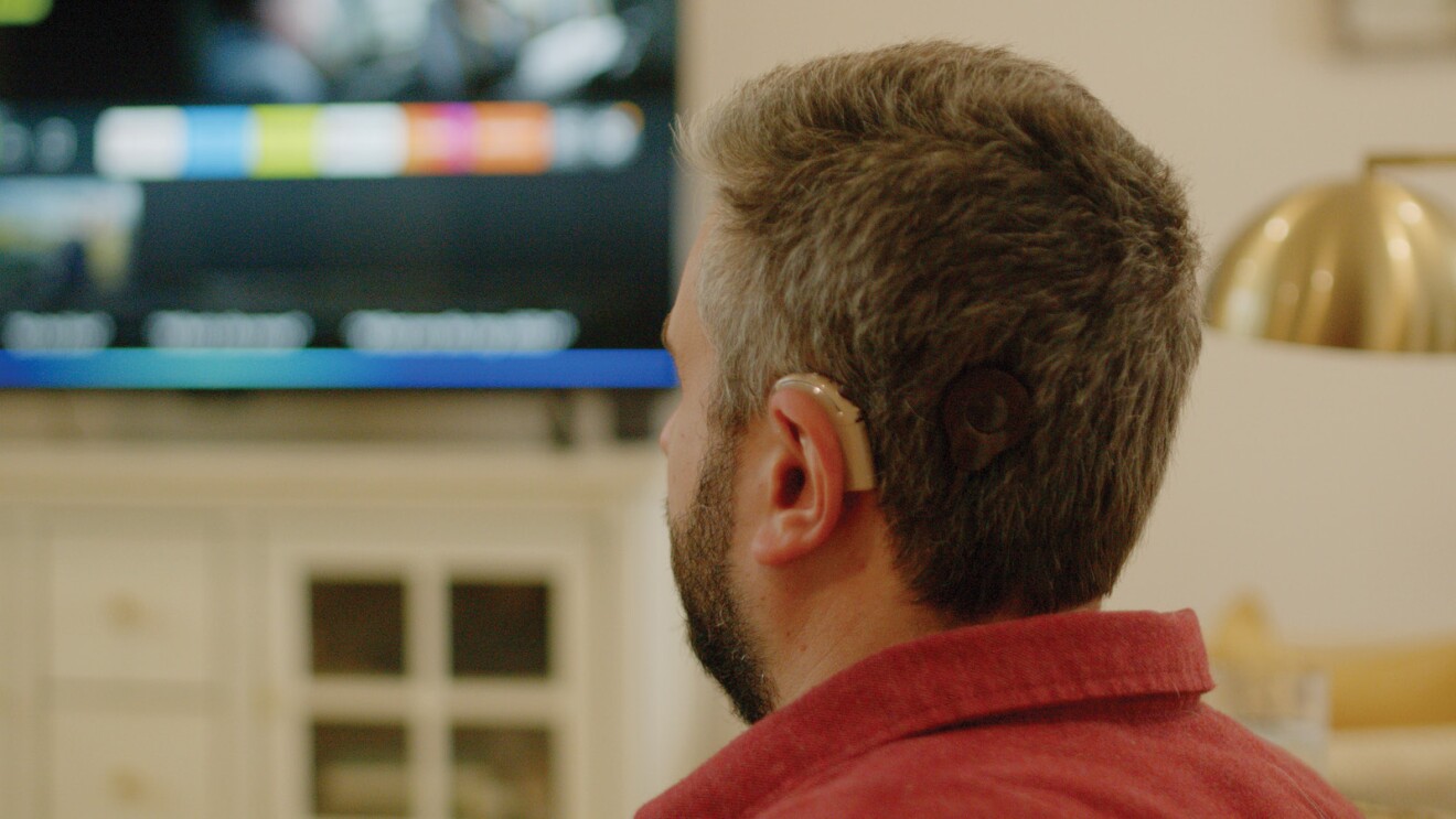 Photograph of Michael Forzano watching TV showing his Cochlear implant in focus in the foreground with the Fire TV in the background.