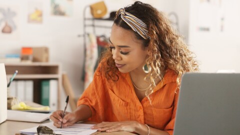 A photo of a business owner writing on a clipboard at a desk. There is also a laptop computer and delivery box next to them.