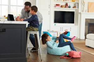 An image of a family interacting with Amazon devices like an Echo Show and a Kindle in their living room. The dad and son are sitting on the kitchen stools and the daughter is sitting on a light blue beanbag with a pink backpack next to her. 