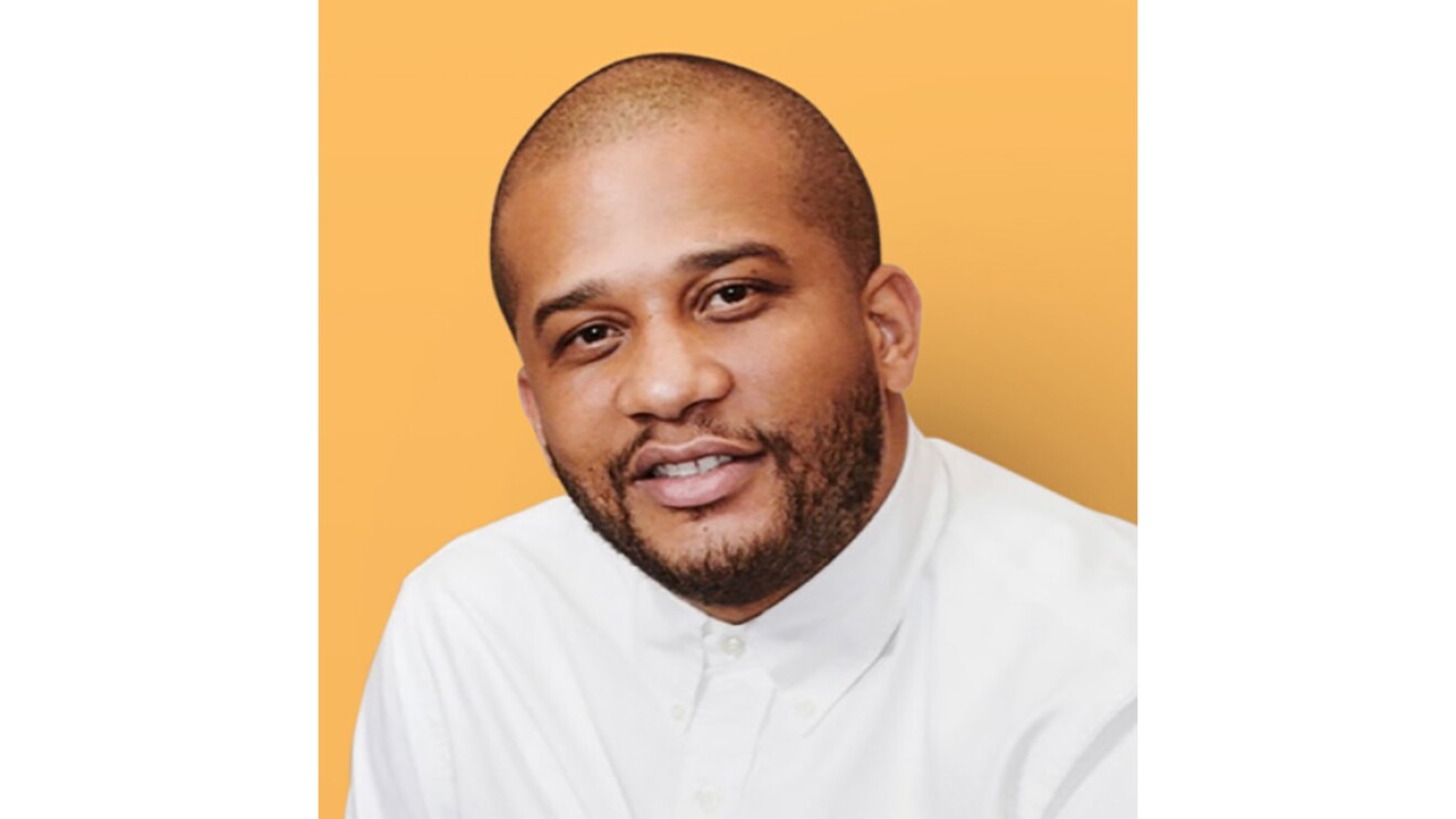A Black man wearing a white button up shirt in front of an orange background.