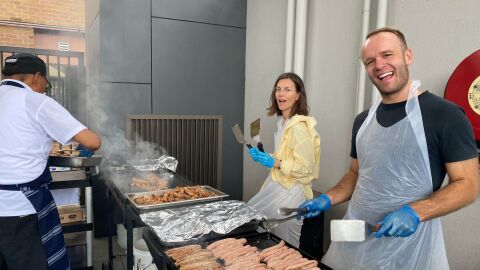 A man in black t-shirt and a a woman in yellow jacket are grilling sausages.