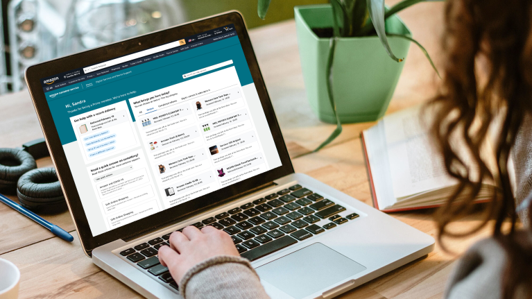 Woman on sitting at a table working on a laptop. On the screen of the laptop is the Amazon Customer Service homepage.