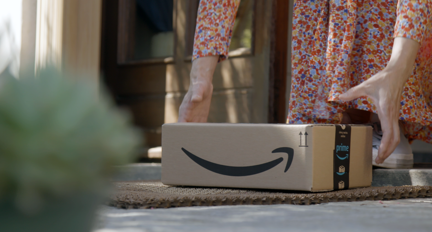 Scatola Amazon in primo piano, posata su uno zerbino di fronte all'ingresso di una porta. Si vedono due mani di una donna che stanno per raccogliere il pacco. La donna indossa una gonna a fiori e delle sneakers. 