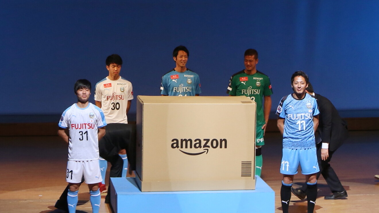 薄暗い室内、スポットライトで照らされたステージに立つ5人の男性。全員ユニフォーム姿で、Amazonロゴの入った巨大なダンボール箱を囲んでいる。