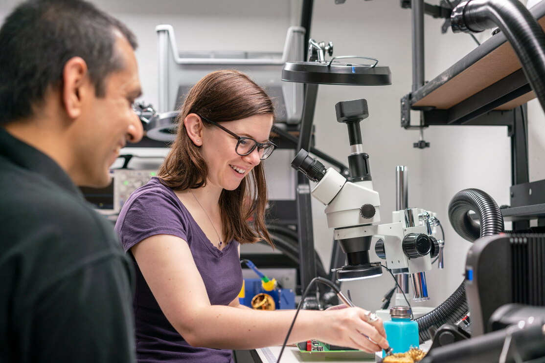 A woman touches the tip of a soldering iron to a sponge, within a laboratory. In front of her sit technical devices, including a microscope. 
