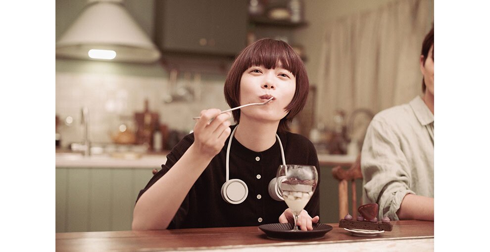 奥にキッチンが見える室内で、テーブルに着席してパフェを食べる女性。首からヘッドフォンを下げている。隣にも人物の半身が見える。