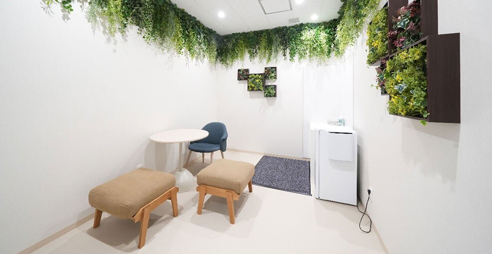 白い壁の小部屋に、丸テーブルと青いチェア、ベージュのスツール2脚が配置。右側には小型冷蔵庫とグレーのマット。天井と壁面にはグリーンが飾られている。