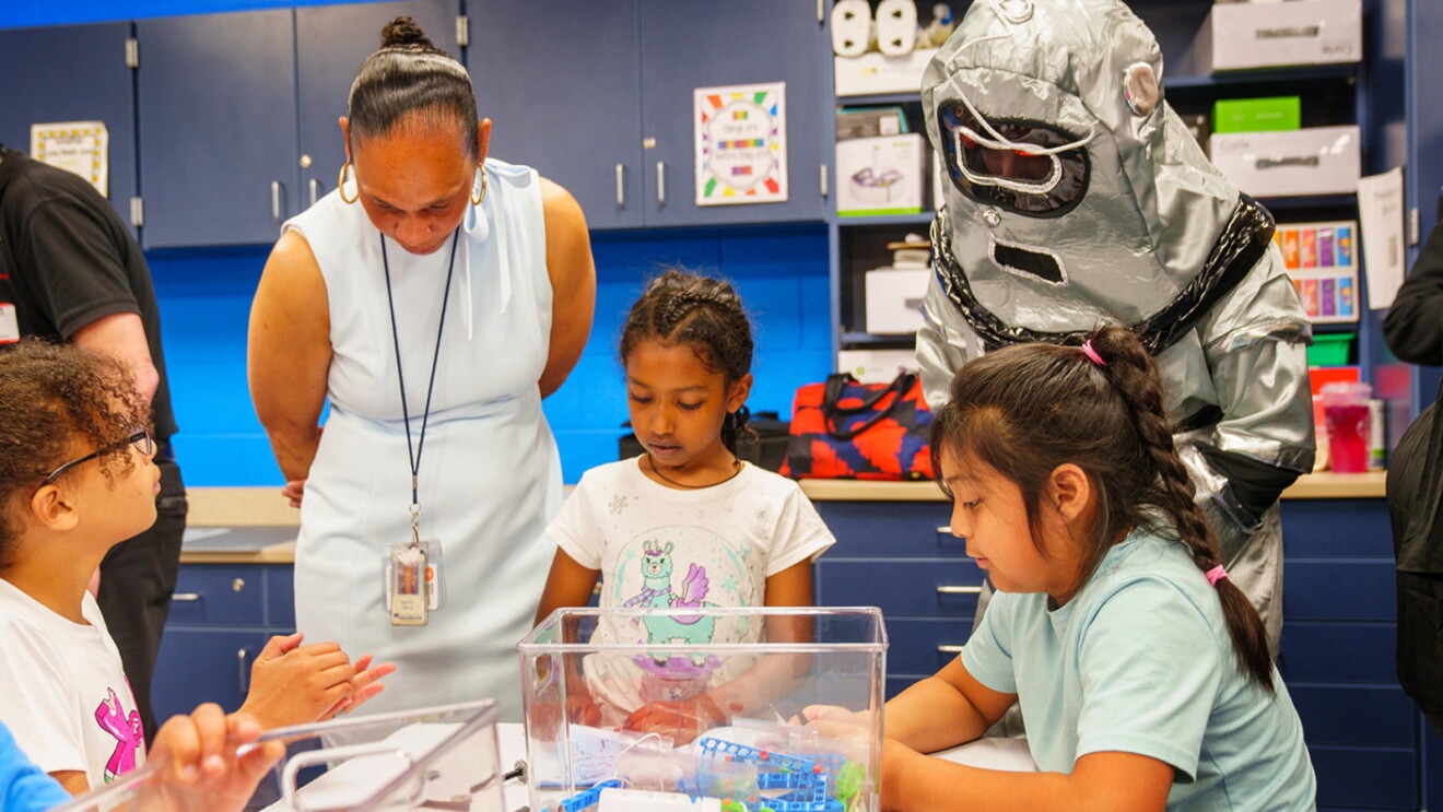 A photo of three students working at a table, with a teacher and astronaut mascot observing, in the AWS Think Big Space at River Oaks Elementary School.