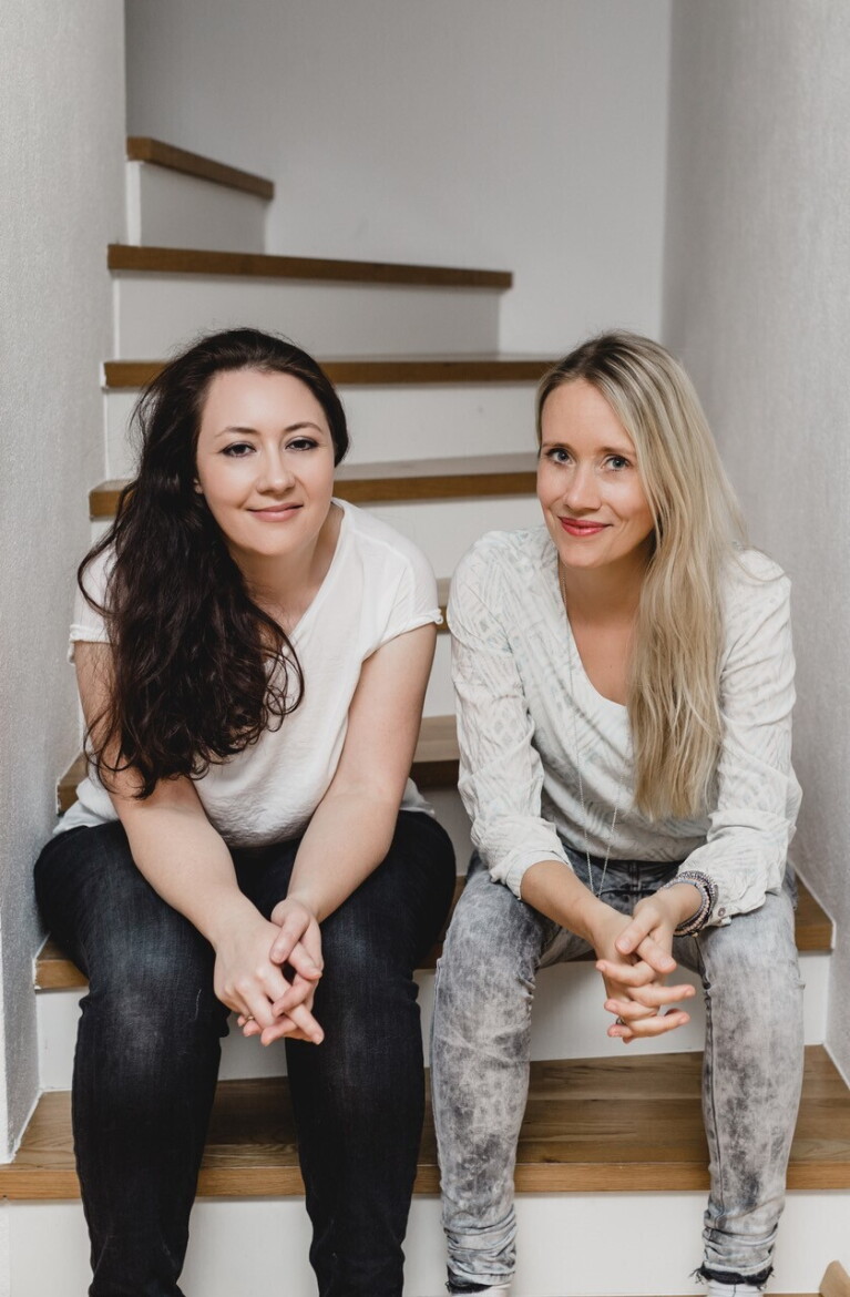 Gruppenfoto des Fantasy-Autorenduos „Rose Snow“, bestehend aus Carmen Schmit und Ulrike Mayrhofer, die beide auf einer Treppe sitzen.