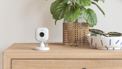 A Blink camera placed on a side table alongside a small plant and a dish containing a pair of sunglasses