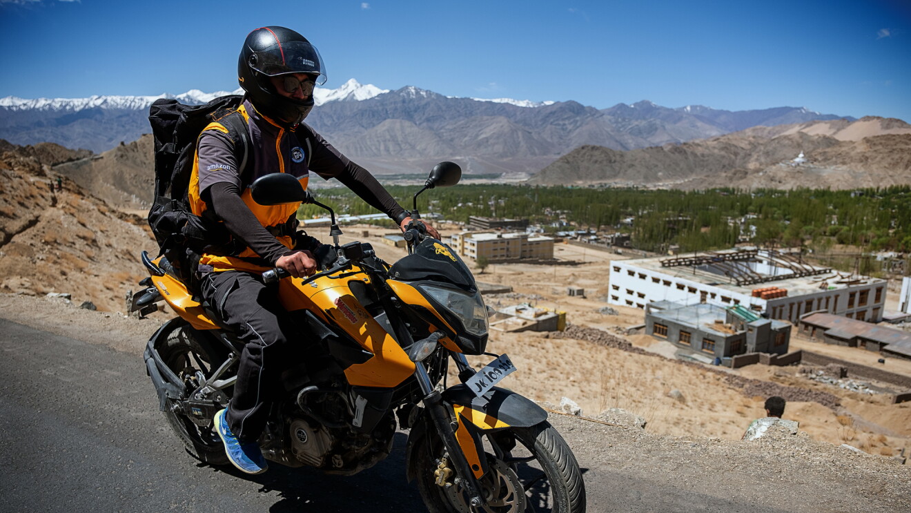 A helmeted motorcyclist wearing a large black backpack rides along an asphalt road. Buildings and snow-capped mountains are in the background of the image.