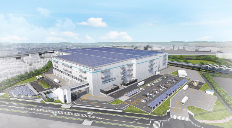 大規模物流施設の空撮イメージ。広大な屋根全面に太陽光パネルを配置。白い建物の側面に「amazon」の文字。周囲には駐車場、ソーラーカーポート、緑地帯、道路が整備され、建物前には大型トラック用の荷捌きエリア。背景には市街地と遠くに山々が広がる。