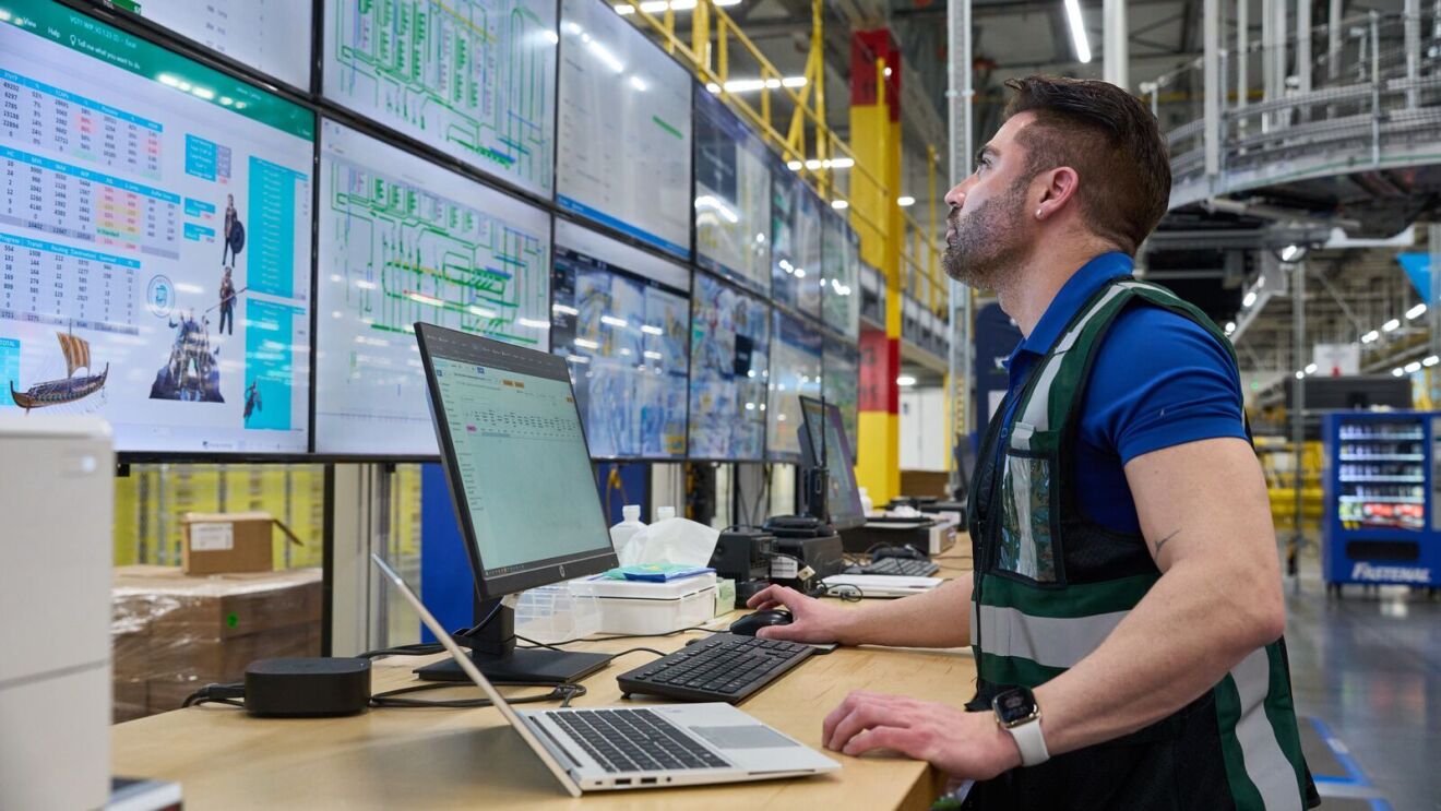 side view of a man looking up at a wall of screens with a keyboard and laptop in front of him inside an amazon fulfillment center