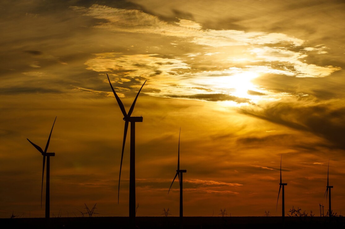 An image of a windfarm in the evening light. Shadows of windmills appear before a sky with an orange sunset and clouds with the sun barely peeking through as it sets.