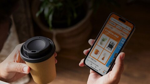 左手に紙製のテイクアウトカップ、右手にスマートホンを持つ人物の手元のショット。スマートホンの画面にはオレンジ色を基調としたAmazonアプリの「おすすめお買い得商品」が表示されている。