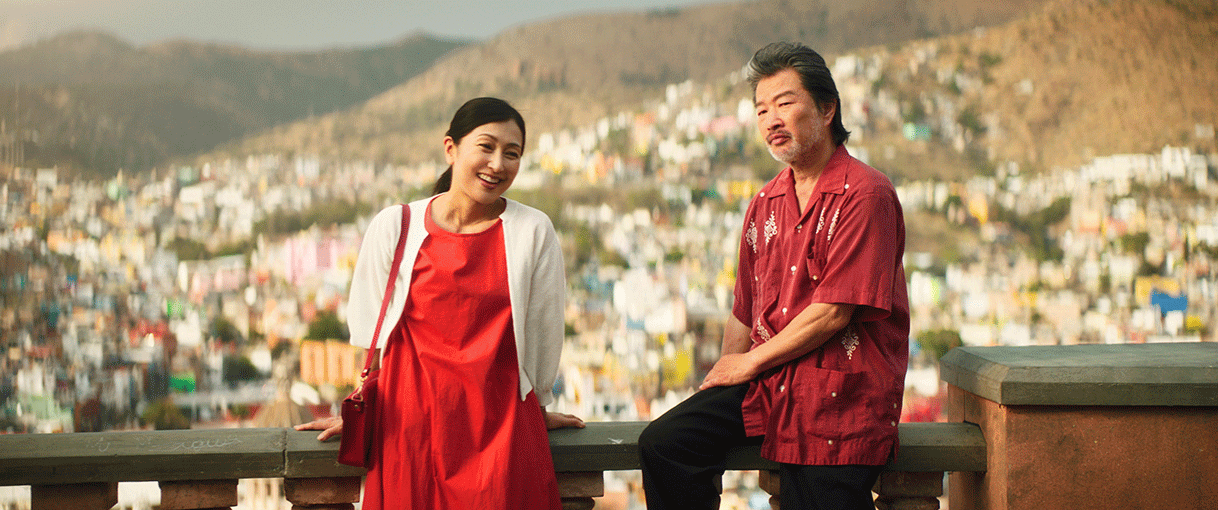 外国のカラフルな街並みを見渡す展望台のような場所で、手すりにもたれた女性と腰掛けた男性。左側の女性は笑顔だが、右側の男性は無表情。