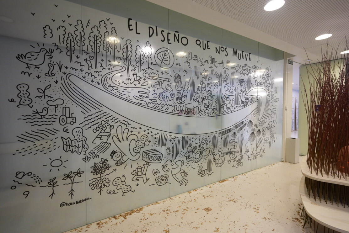 Sobre una pared de cristal se ha pegado un vinilo con un mural. En el centro está la sonrisa de Amazon, y a su alrededor se distribuyen distintos dibujos de personajes haciendo cosas en un entorno natural, con árboles y montañas. 