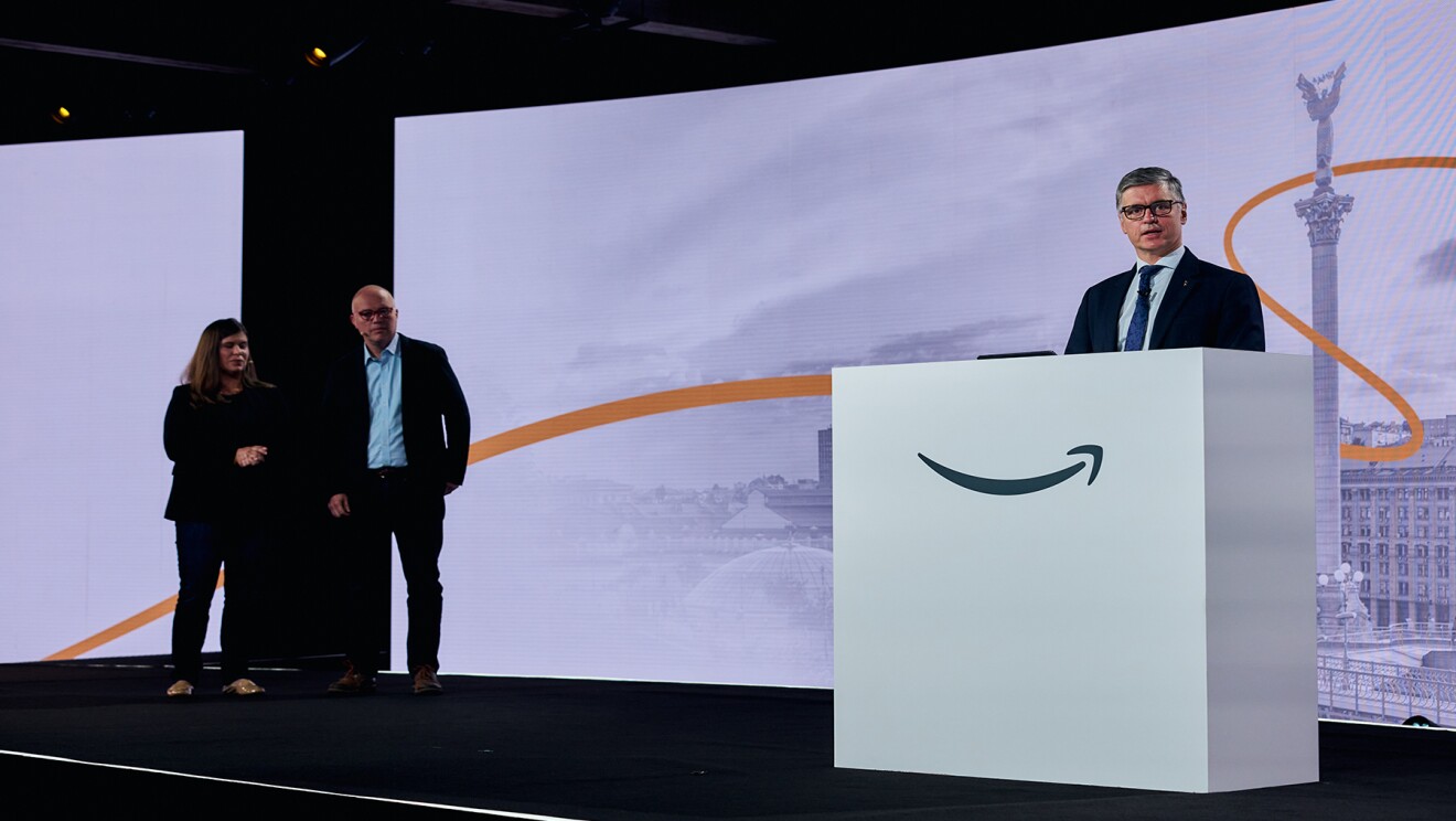 Ukraine’s Ambassador to the UK Vadym Prystaiko stands behind a white podium with the Amazon smile logo on it as he addresses Amazon employees. Liam Maxwell, director of the Government Transformation team in the AWS public sector, and AWS Director of Global Social Impact Maggie Carter stand on the stage listening to Prystaiko speak.
