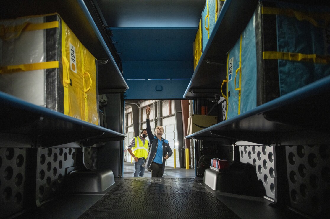 Two people stand at the back of a delivery vehicle and look at the shelving designed to hold Amazon customer orders.