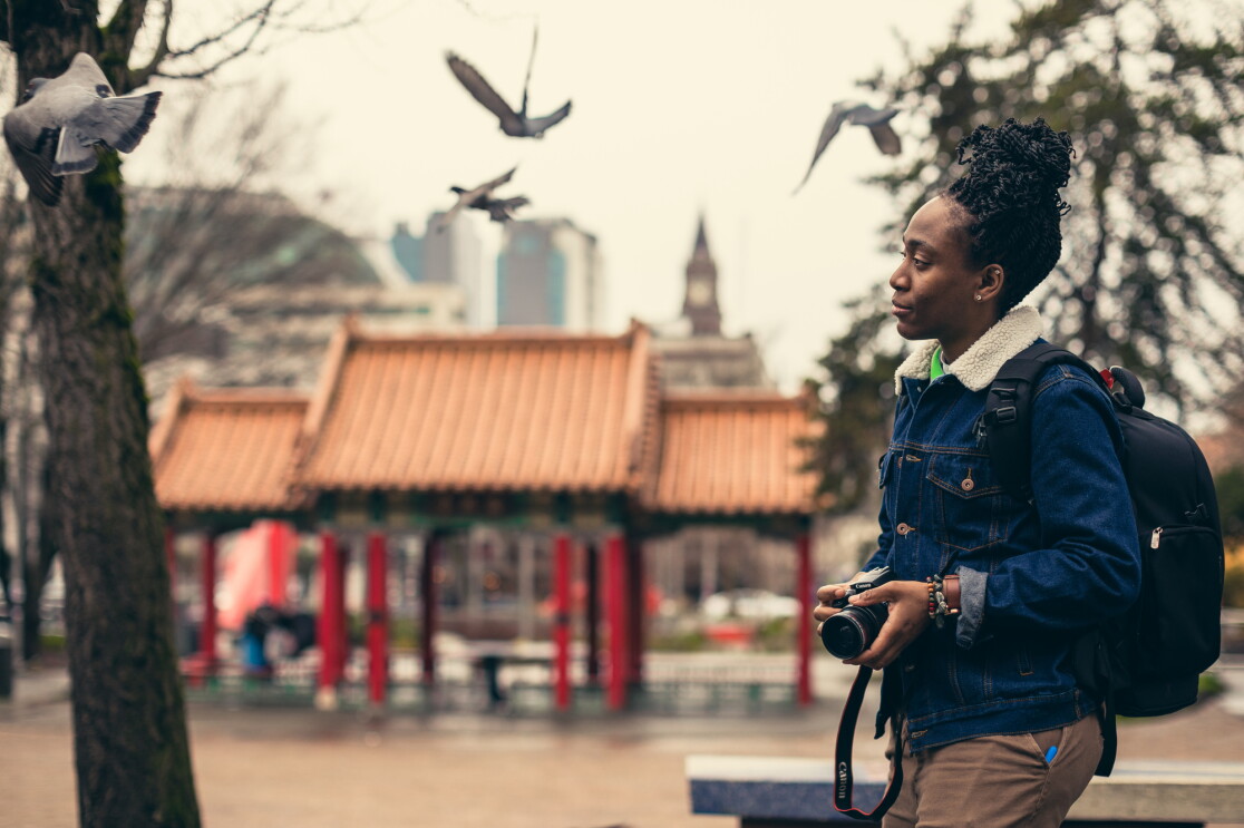 A woman in a blue jacket, green top, and brown pants walks in front of a temple. The Seattle skyline is behind her. 