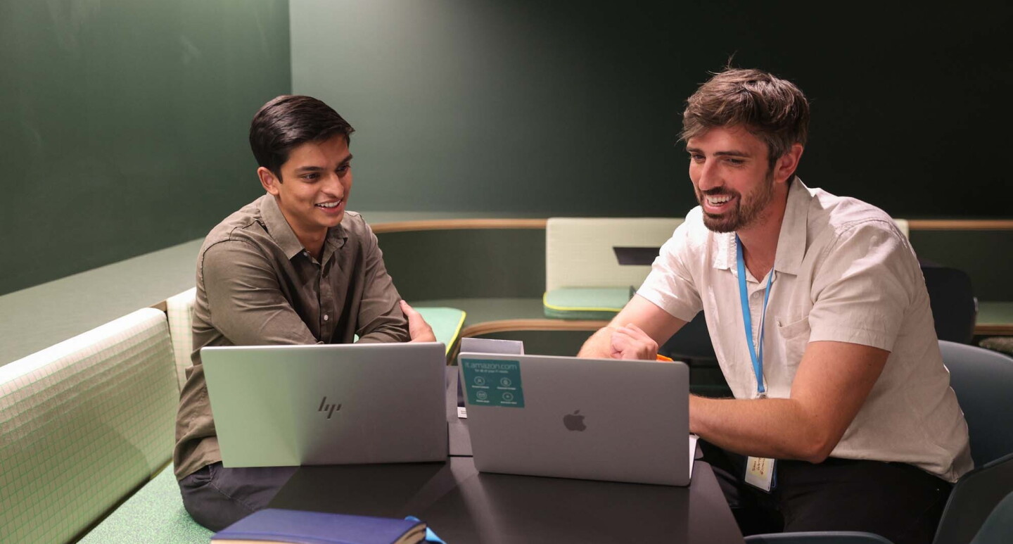 Two men, who are Amazon employees, are sitting in a shared work area at our Sydney, Australia office, working together on their laptop computers. 