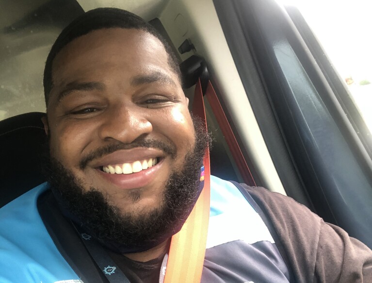 An image of a man smiling for a selfie in his car. He is wearing the blue Amazon delivery vest.