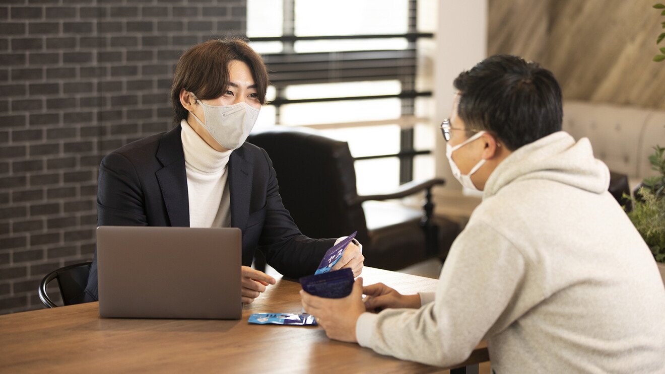 オフィスでテーブルをはさんで向き合って座る男性2人。手に商品をもって話し合っている