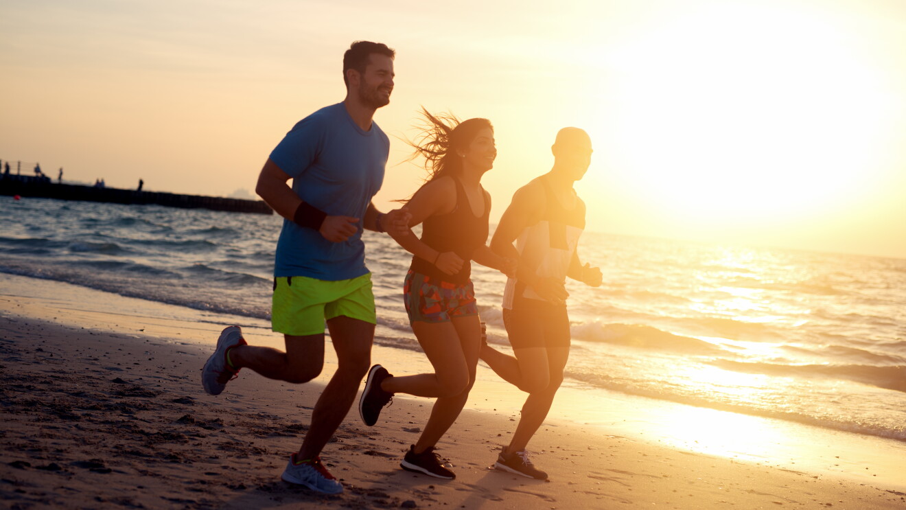 Two men and a woman run on a beach at sundown.