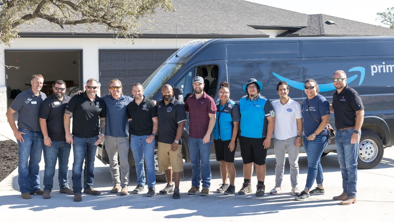 A group of Amazon employees and members of the Gary Sinise Foundation standing side-by-side in front of a smart home and Amazon Prime van.