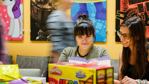 Two women in the foreground sit at a table surrounded by packaging. Other people pass by in the background. The wall has posters of My Little Pony, Play-Doh, and other Hasbro brands.