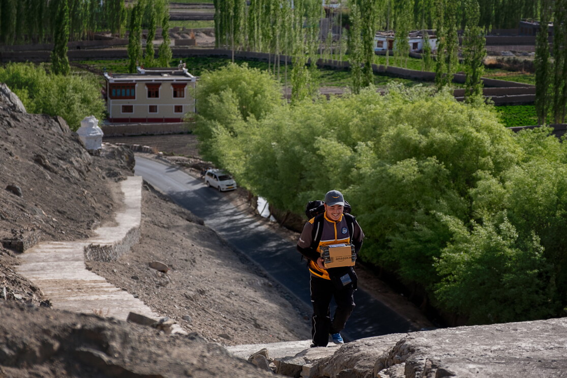 A smiling man in an Amazon delivery uniform carries a package up a staircase of at least 100 stairs that snakes along a hillside up from a road below. Trees and buildings are in the background of the image.